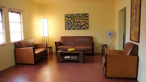A living room with a brown leather sofa, two brown armchairs, a coffee table with a bowl of fruit, a floor lamp, and a painting on the wall.