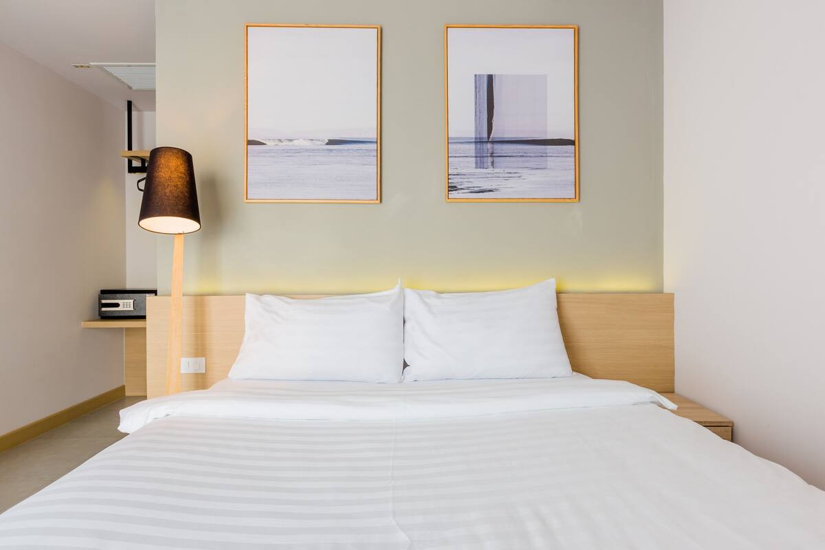 A hotel room with a bed, a bedside lamp, two framed pictures on the wall, and a small shelf with a radio.