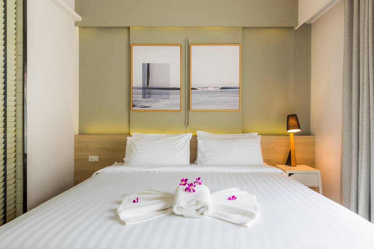A hotel room with a large bed, two white pillows, a nightstand with a lamp, and a view of the ocean through a window.