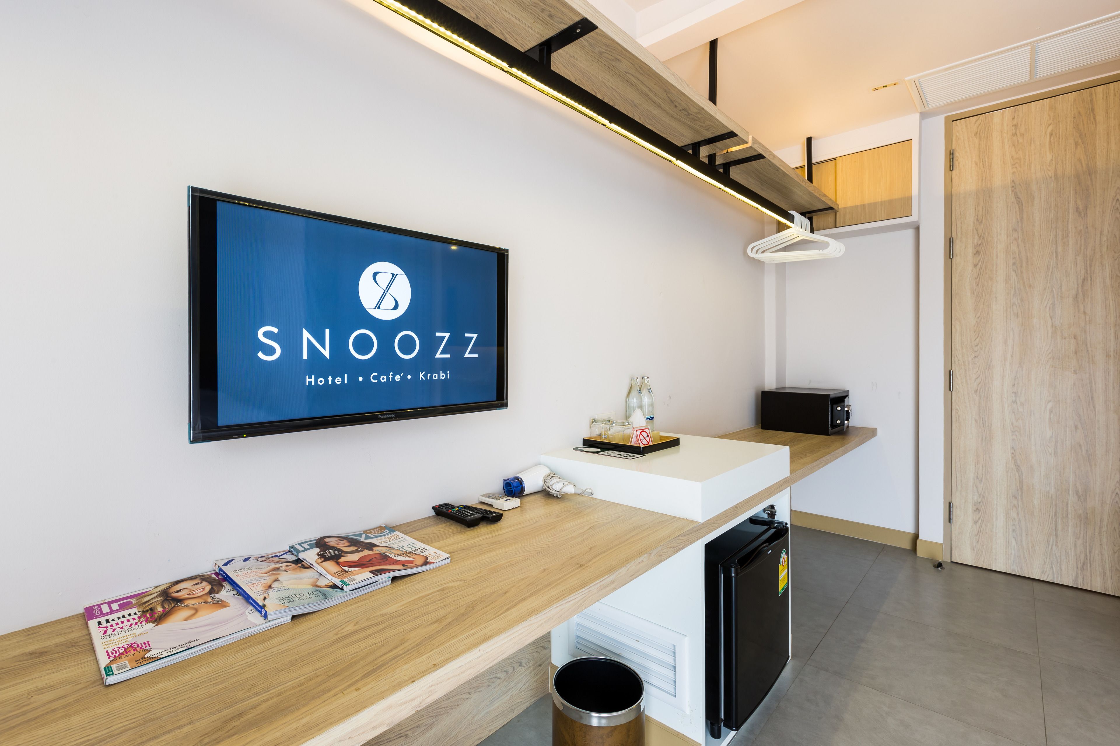 A reception desk with a flat-screen TV displaying the hotel's name, a wooden counter, a trash bin, and a small refrigerator.