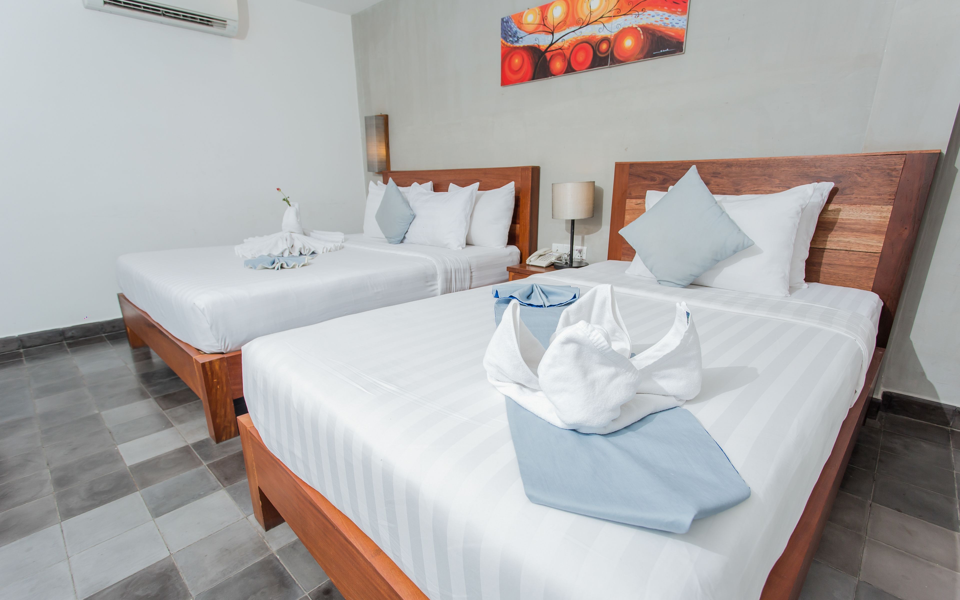 Ein Hotelzimmer mit zwei Betten, jeweils mit weißer Bettwäsche und einem hölzernen Kopfteil. Darüber hängt ein Bild an der Wand und auf dem Nachttisch steht eine Lampe.
