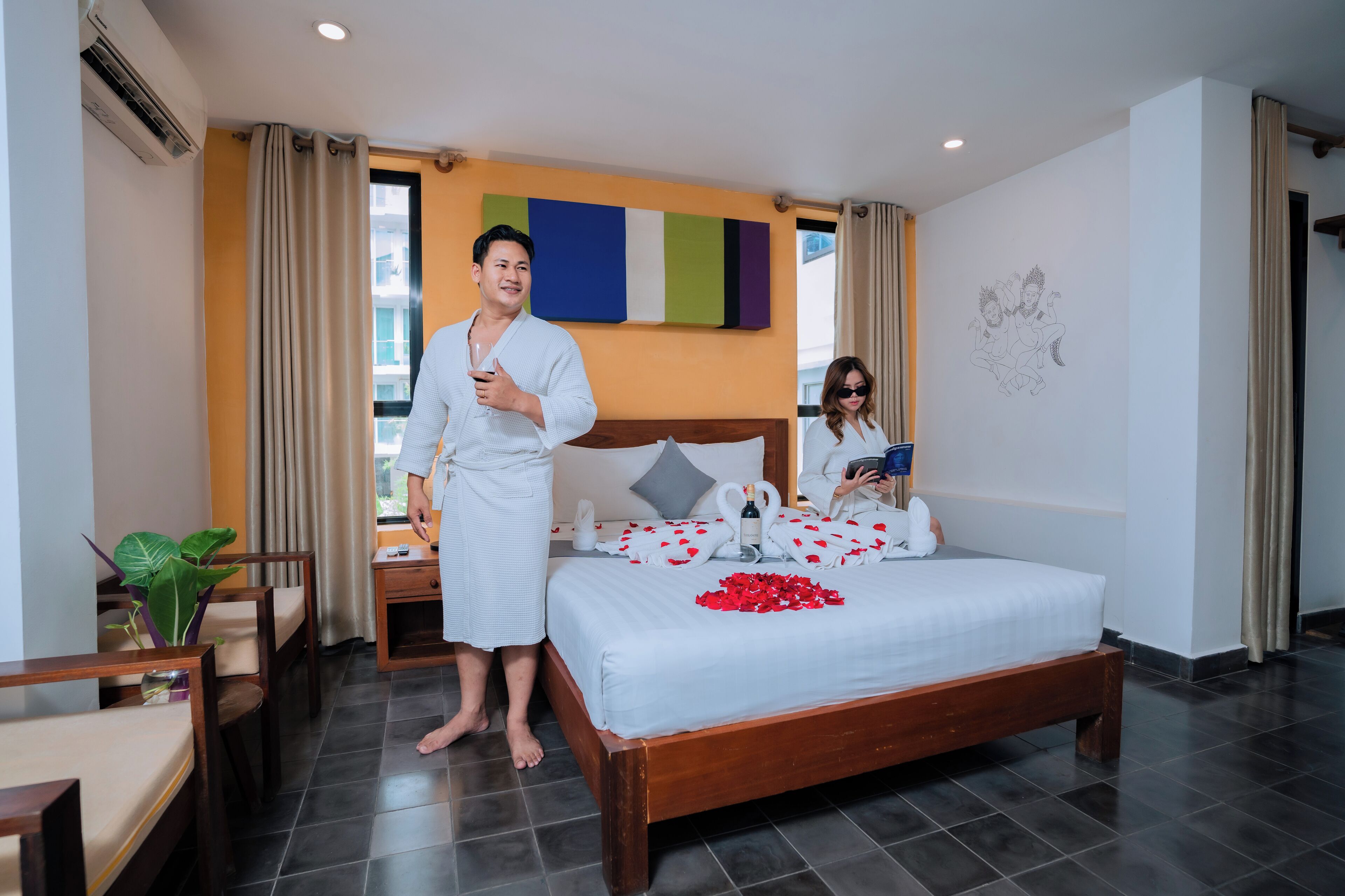A hotel room with a bed, a bedside table, a nightstand, a potted plant, and a window with curtains.