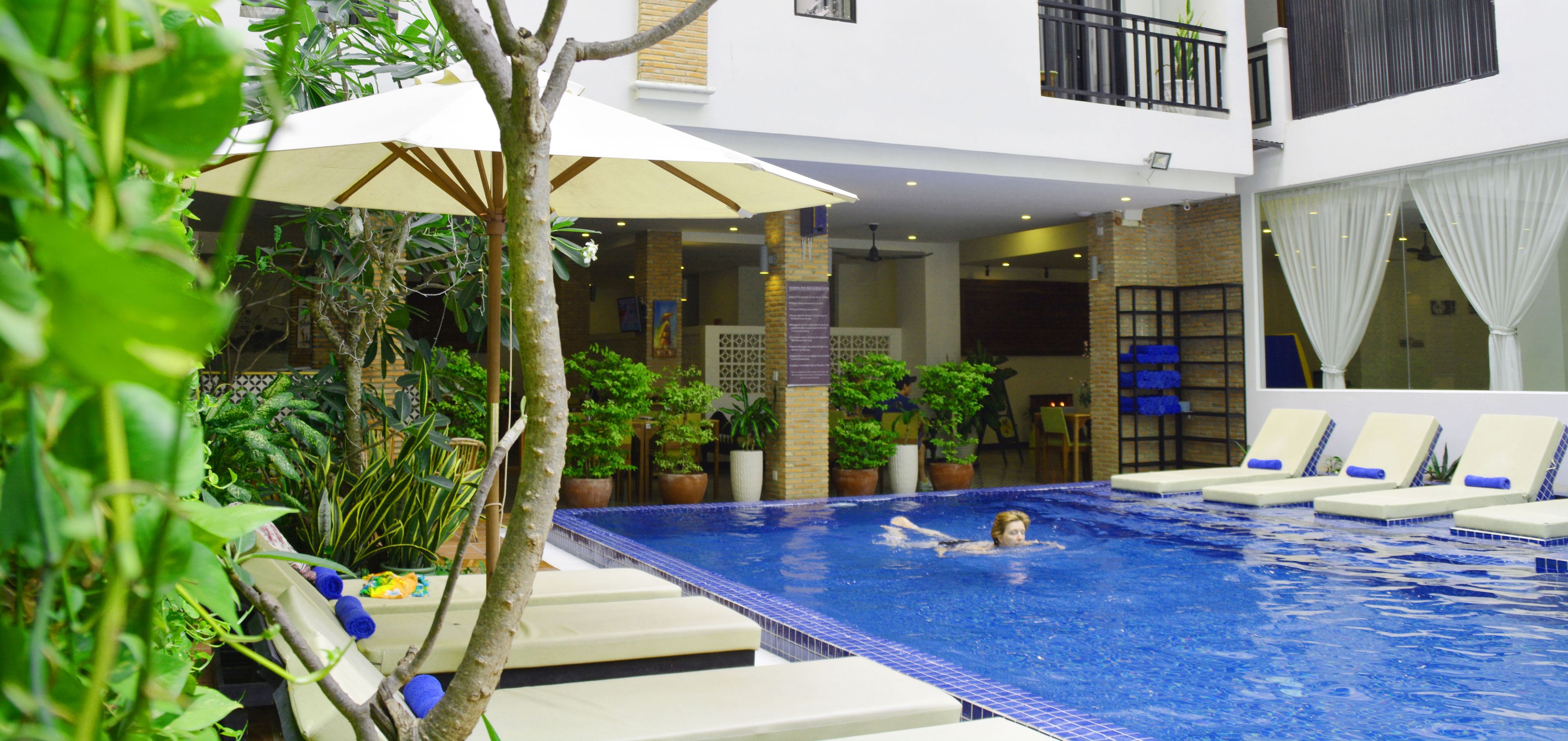 A swimming pool with lounge chairs and umbrellas, surrounded by potted plants and a building with a balcony.