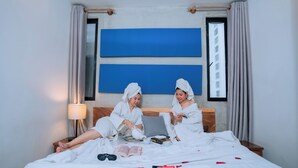 Et hotelværelse med en seng, to personer der læser, en lampe og et blåt kunstværk på væggen.