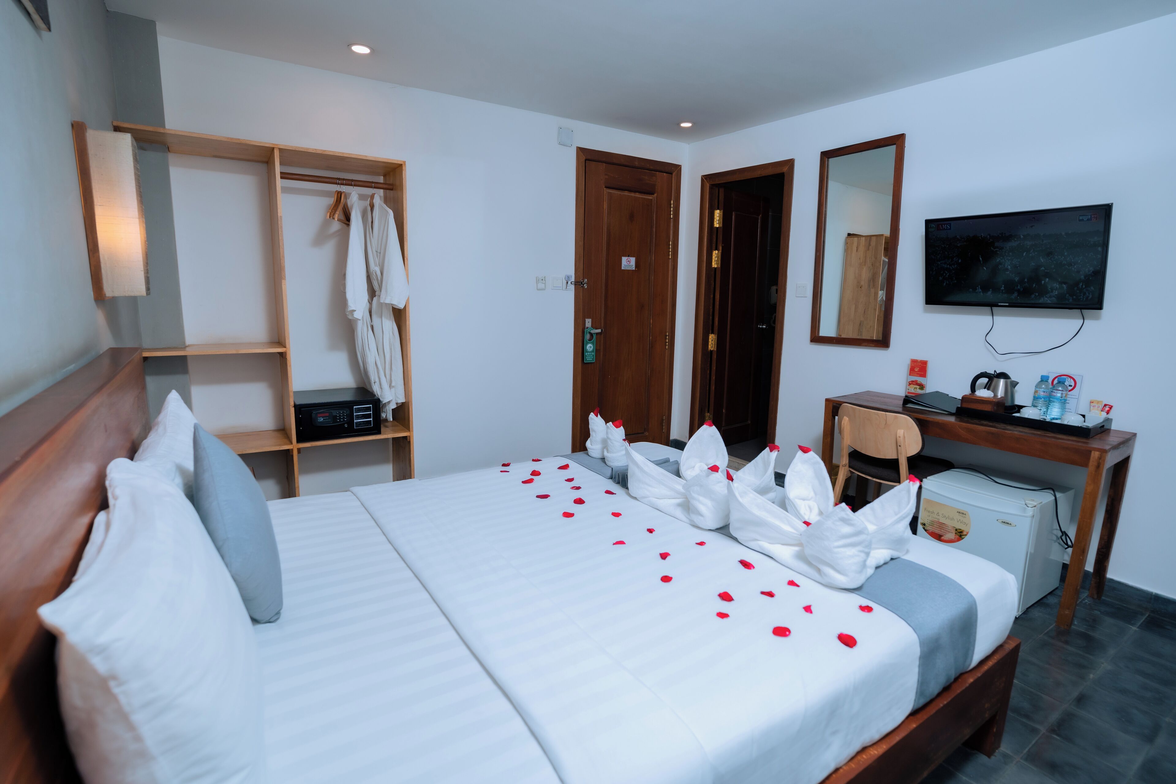 A hotel room with a neatly made bed, a wardrobe, a desk with a chair, a television, and a refrigerator.