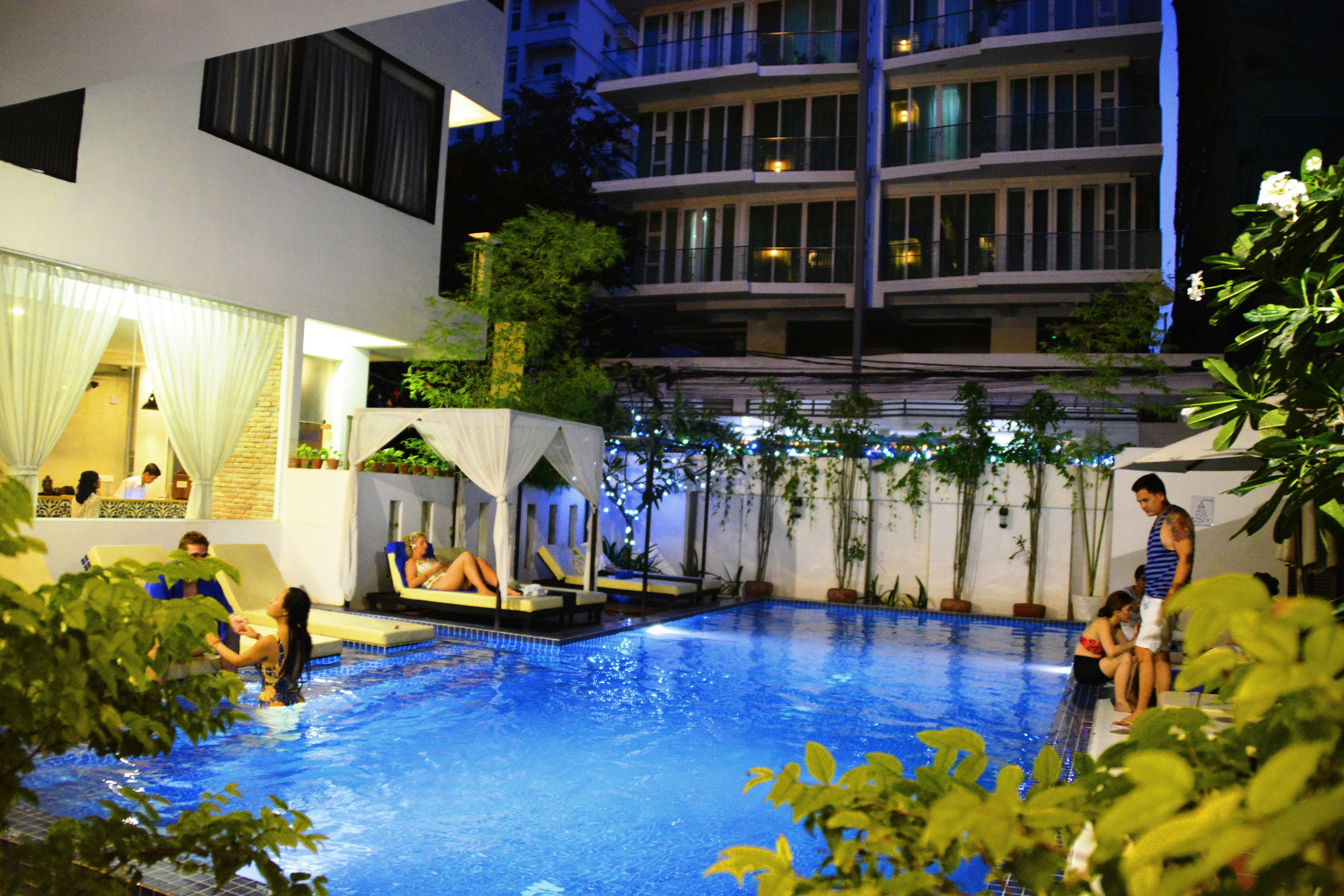 An indoor swimming pool with people relaxing and sunbathing. There are potted plants around the pool area and a building with balconies in the background.