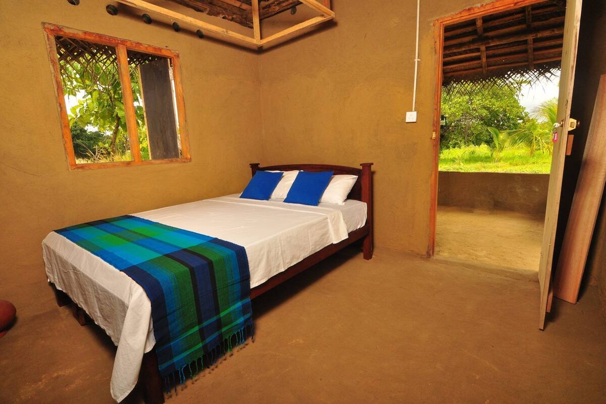 A single bed with a white mattress and blue and green striped blanket, wooden headboard, and a window with a view of greenery.