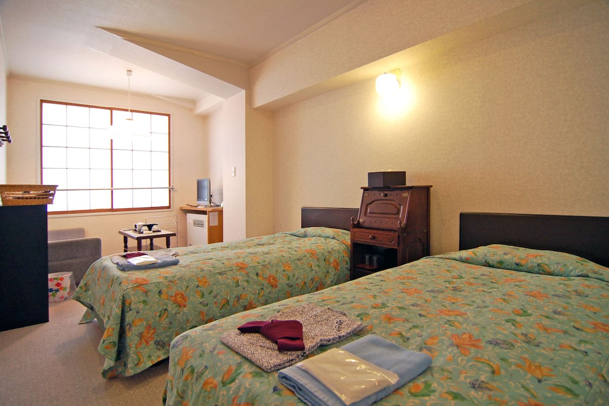 A hotel room with two beds, a desk, a chair, and a window.