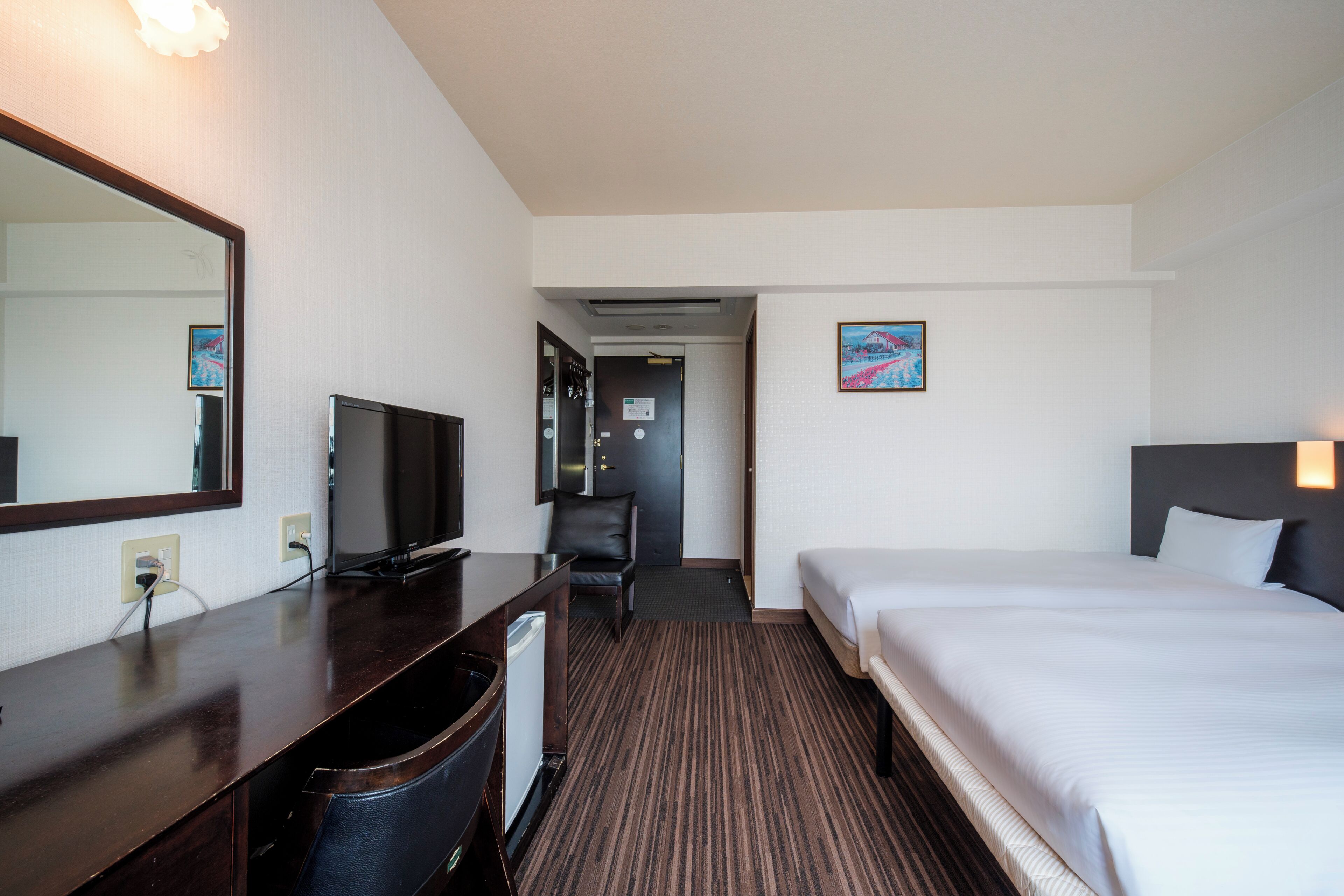 A hotel room with a bed, a desk with a television, a chair, and a door leading to a hallway.