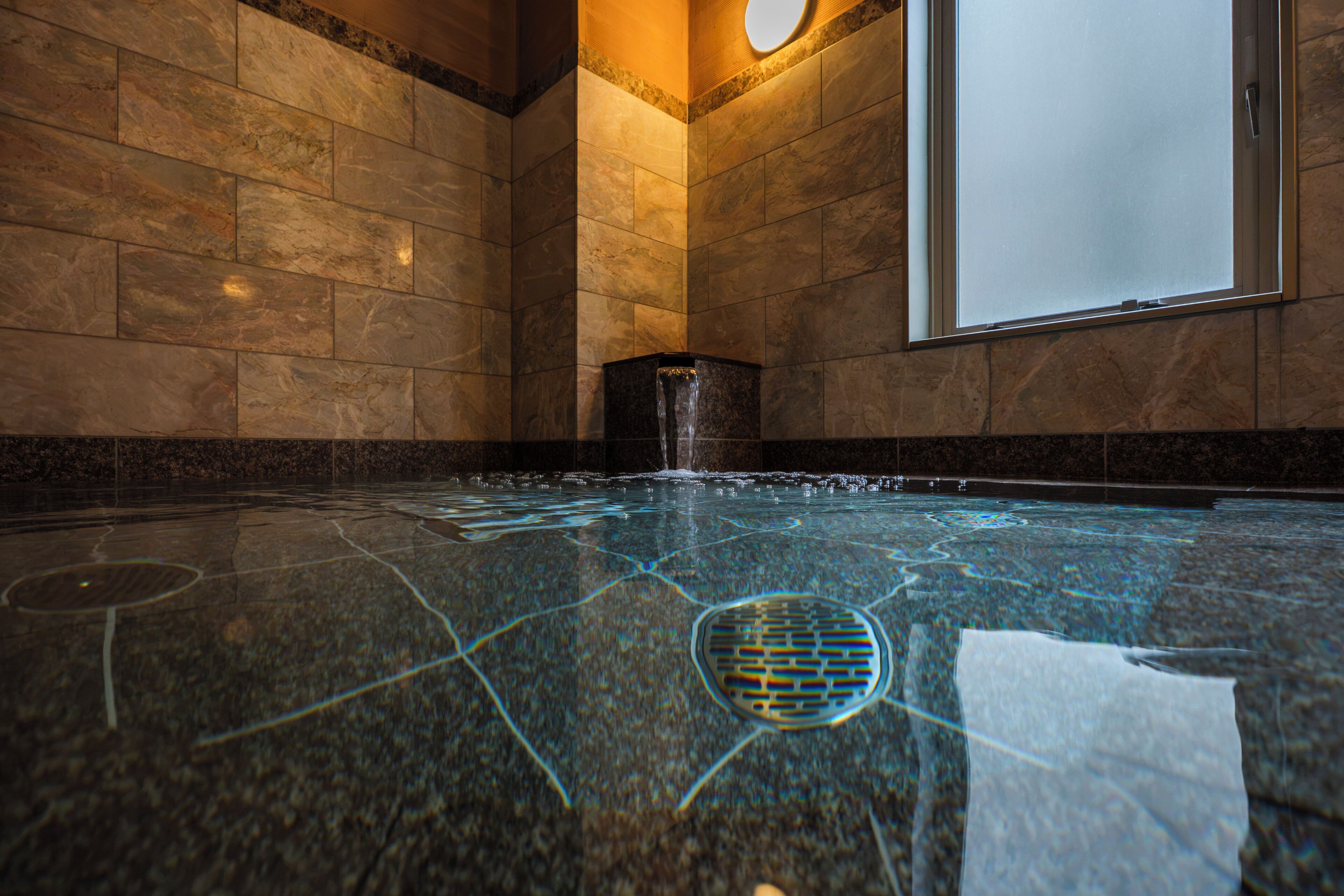 Une piscine intérieure moderne, dotée d’un carrelage aux motifs géométriques, d’un élément décoratif aquatique et d’une grande fenêtre.