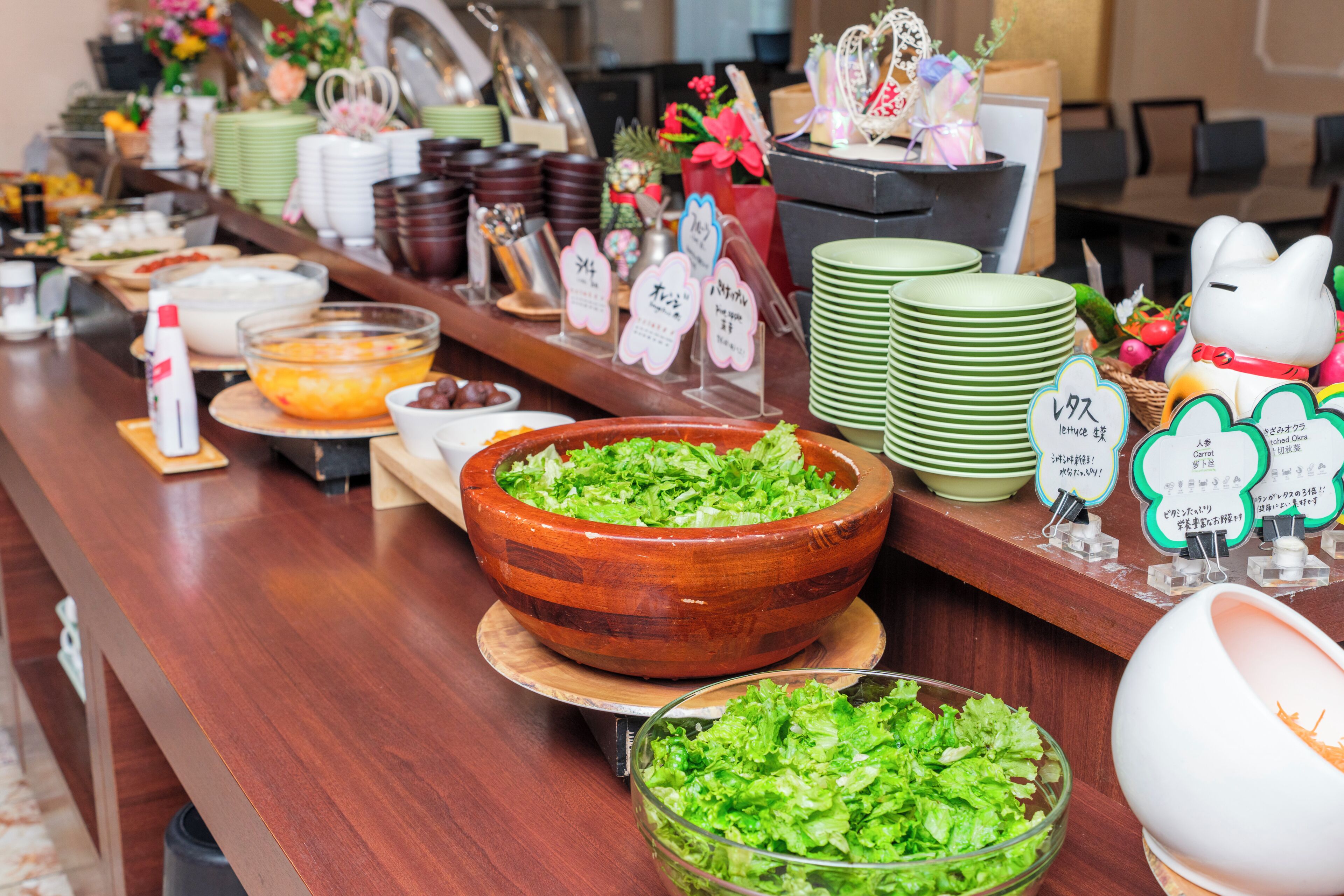 A buffet table with various dishes, including a large wooden bowl of salad, green plates stacked beside, and signs indicating food options.