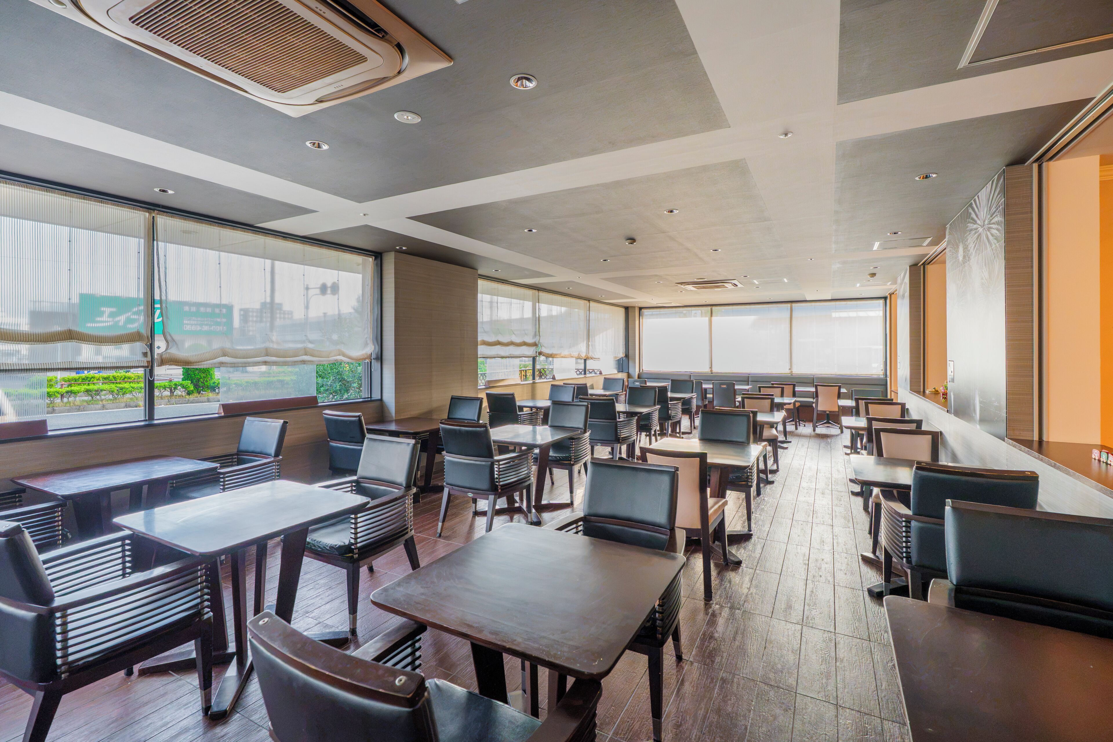 A spacious indoor area with multiple tables and chairs, large windows offering views of the outside, and a modern ceiling design.
