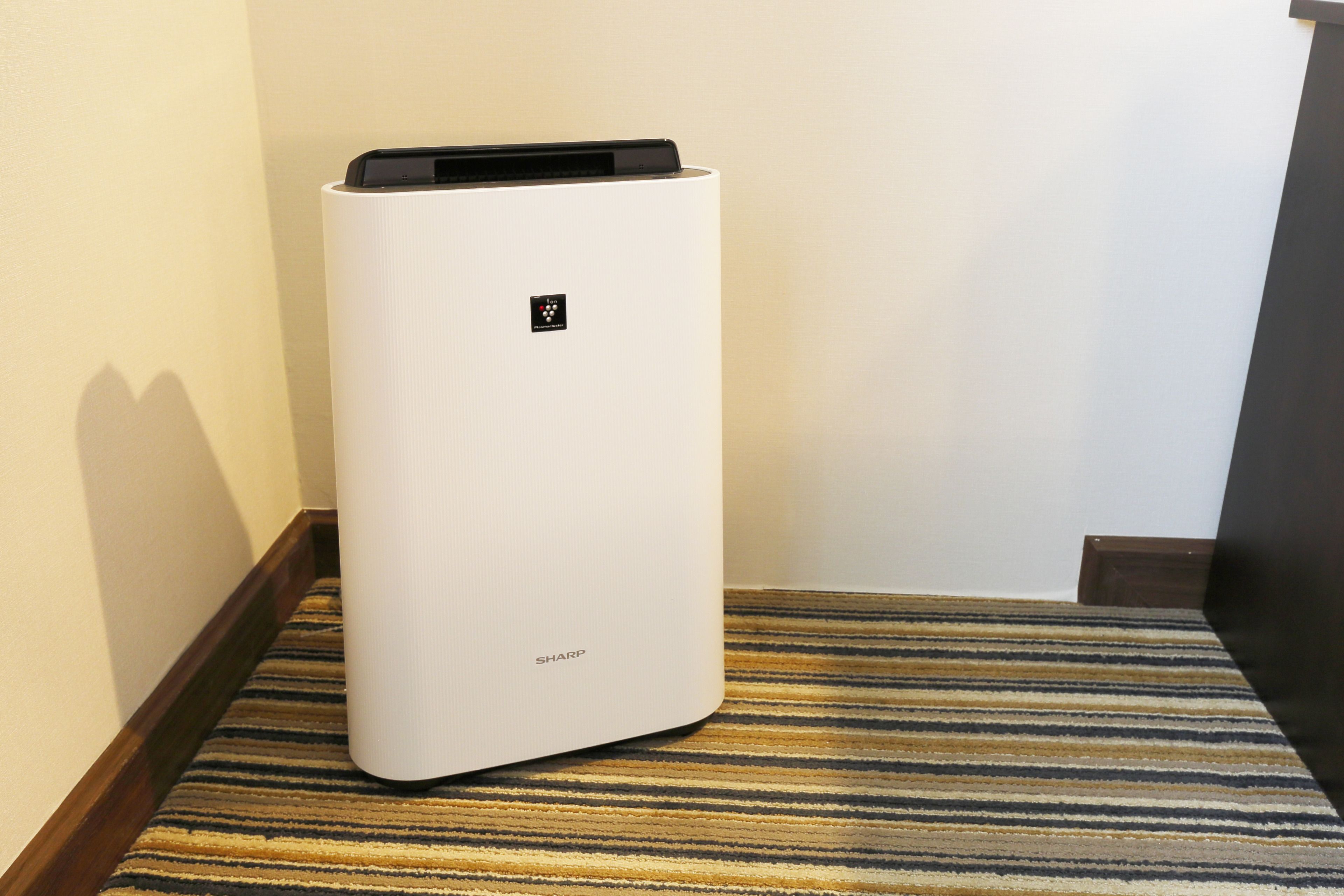 A white air purifier with a control panel on top, placed on a striped carpet in a corner of a room.