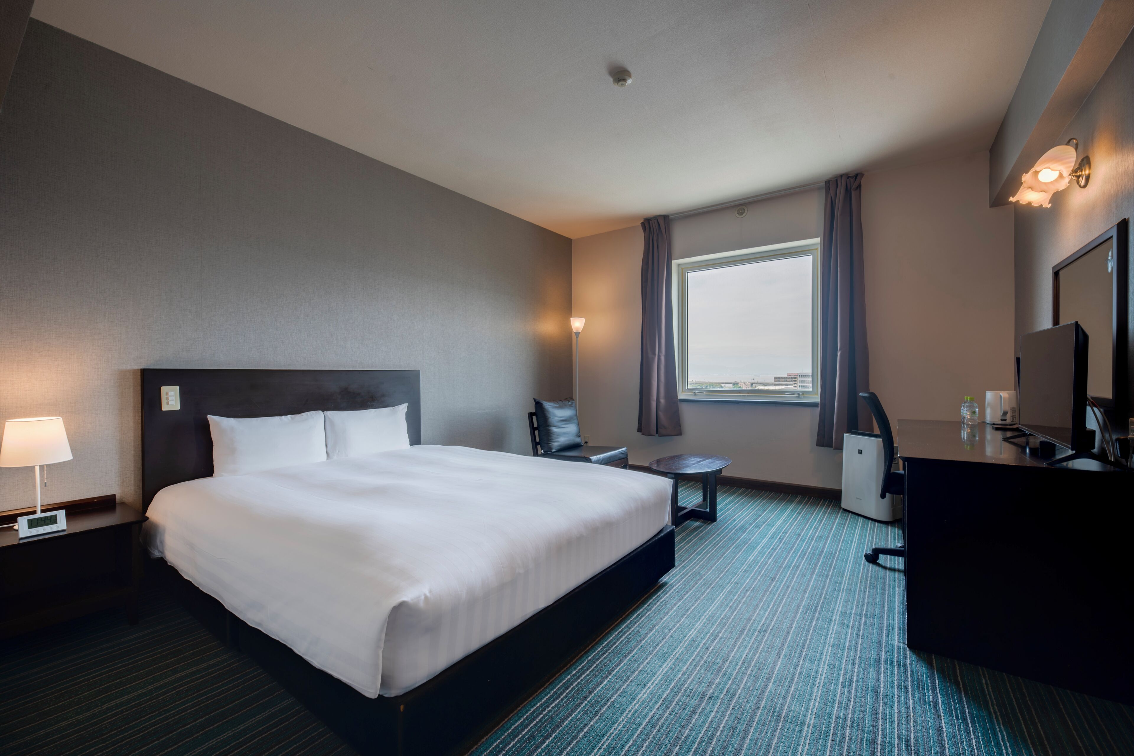 Habitación de hotel con una cama grande, un escritorio, una silla, una mesita, una lámpara de escritorio, una cafetera, un espejo y una ventana con cortinas.