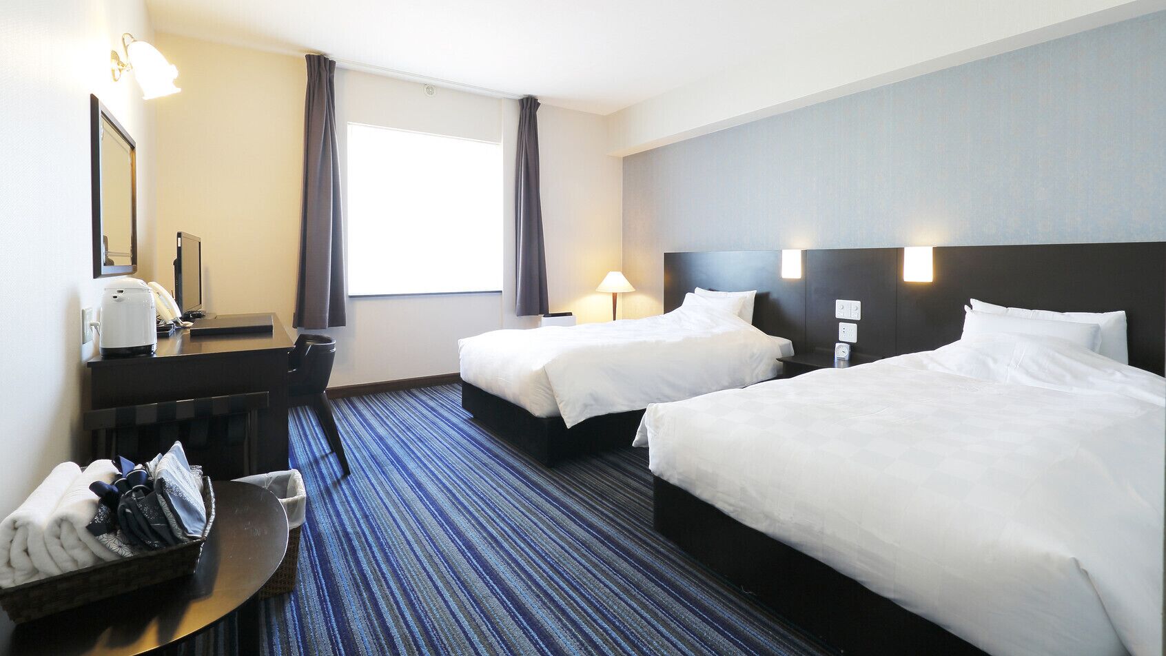 A hotel room with two beds, a desk with a chair, a desk lamp, a bedside table, and a window with curtains.