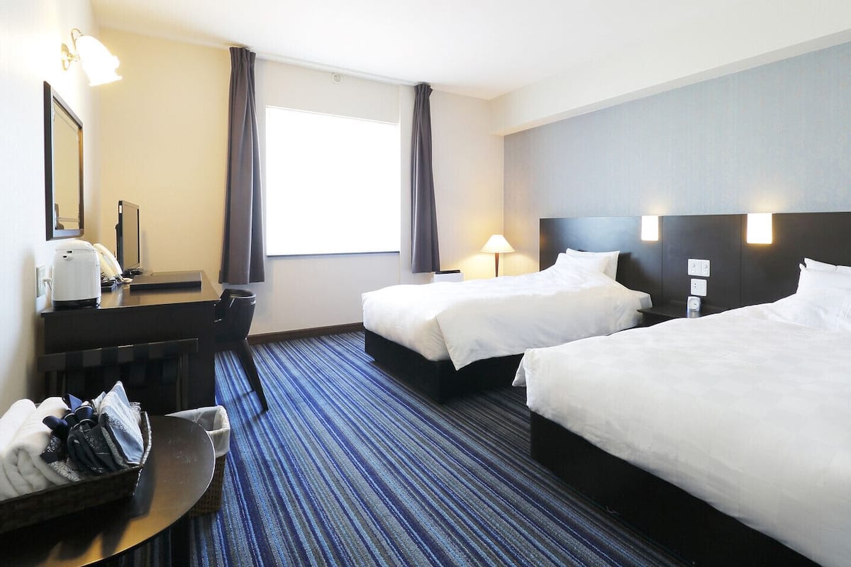A hotel room with two beds, a desk with a chair, a desk lamp, a bedside table, and a window with curtains.