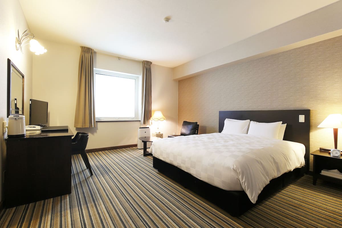 A hotel room with a large bed, a desk with a computer, a chair, a lamp, and a window with curtains.