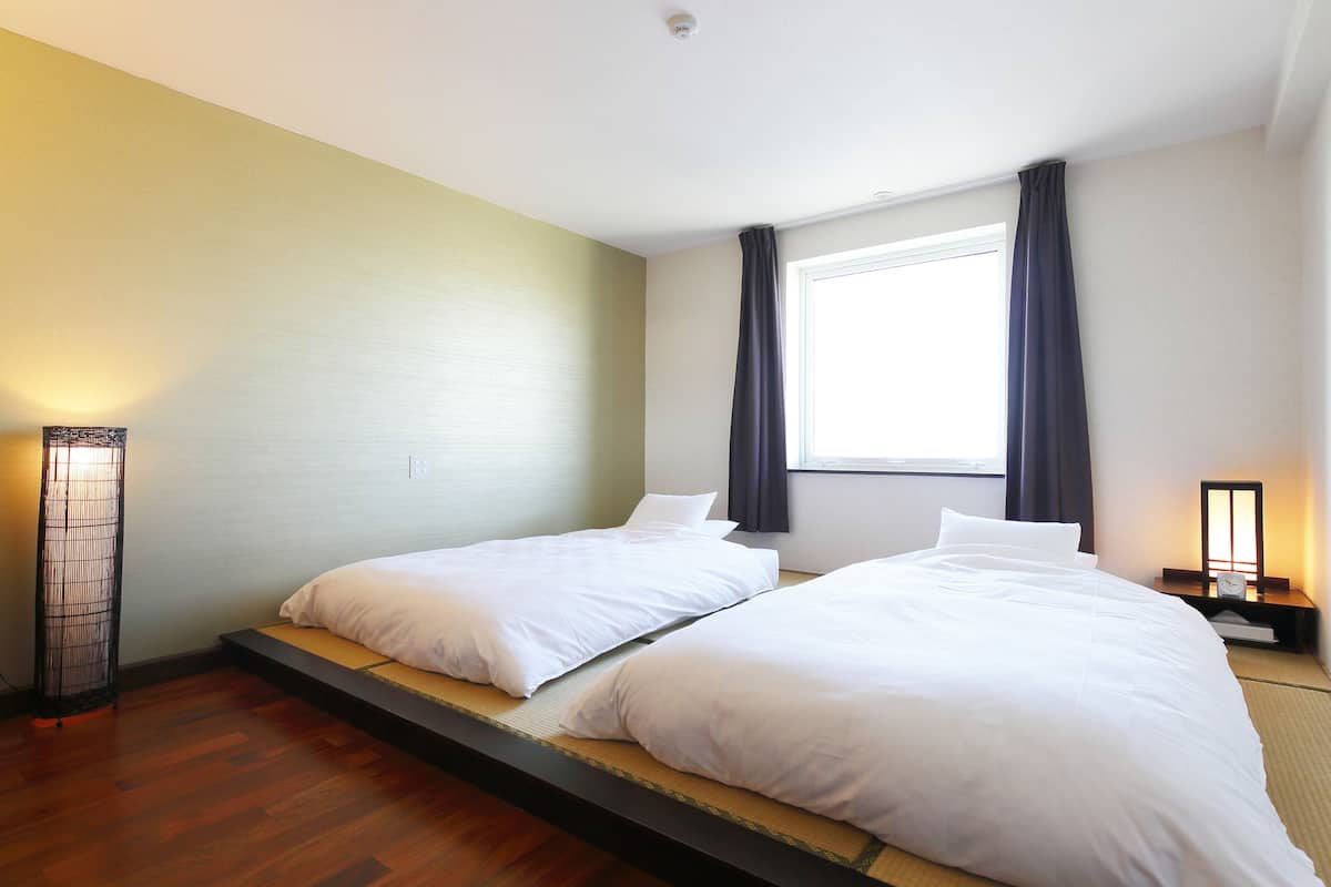 A hotel room with two single beds, a standing lamp, a nightstand with a clock, and a window with curtains.