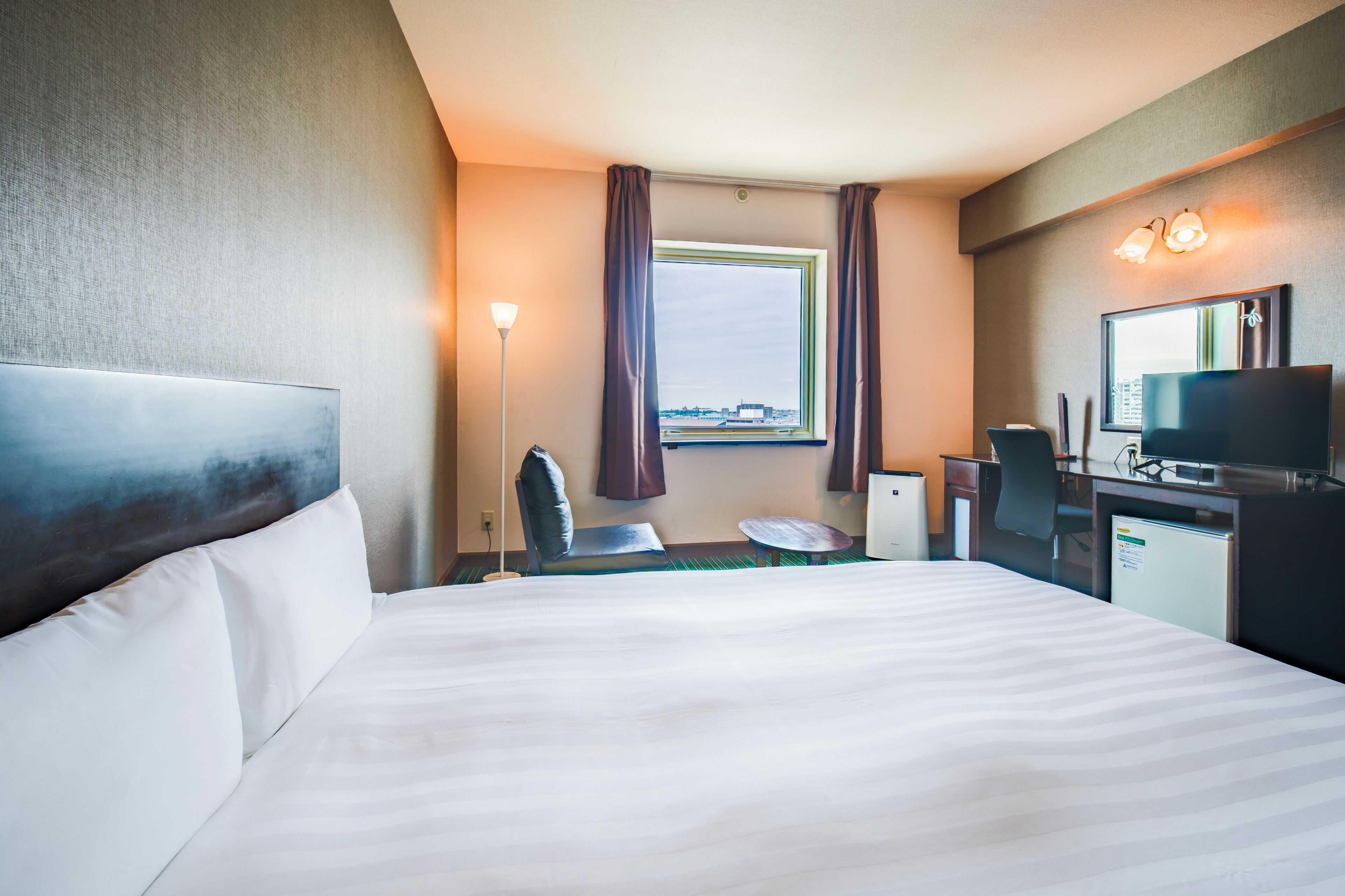 A hotel room with a bed, a desk with a computer, a chair, a small table, and a window with curtains.