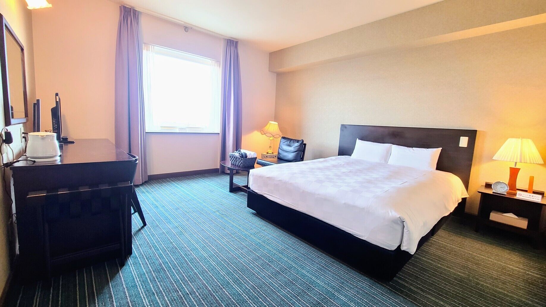 A hotel room with a bed, a desk, a chair, a lamp, and a window with curtains.