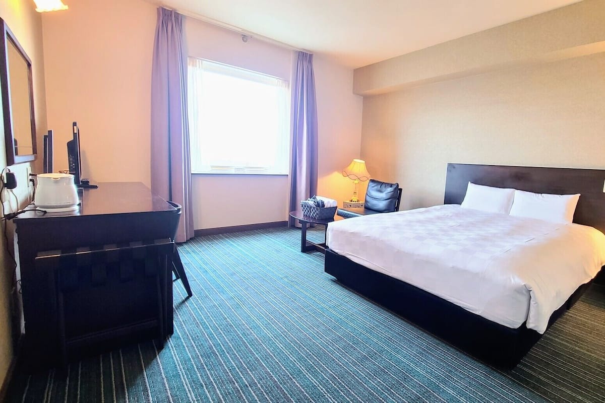 A hotel room with a bed, a desk, a chair, a lamp, and a window with curtains.