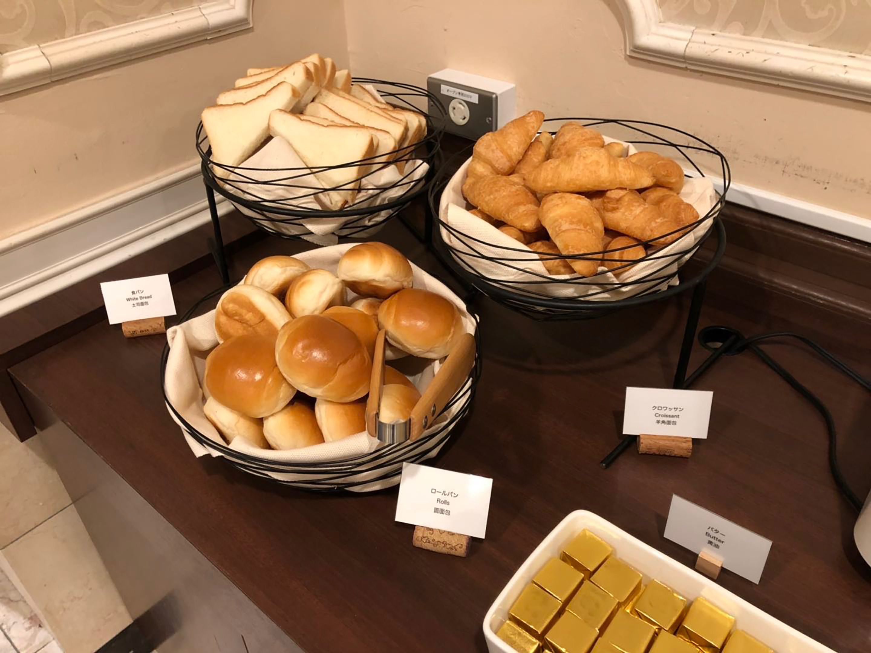 A buffet table with labeled food items including white bread, croissants, rolls, and butter.