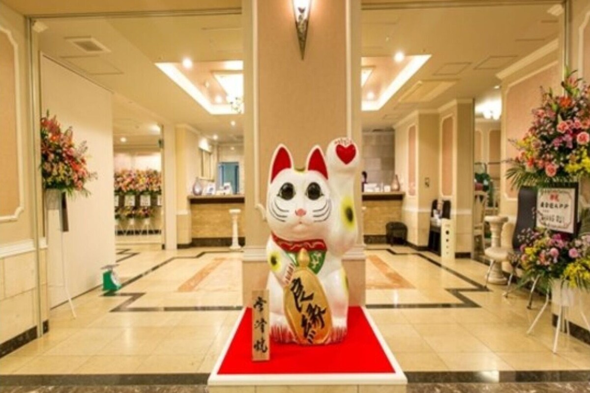 A hotel lobby with a decorative cat statue on a red platform, a reception desk, and floral arrangements.