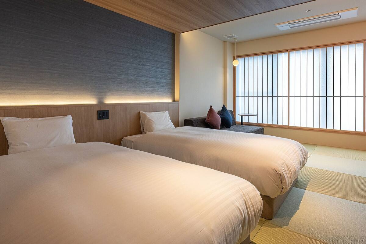 A hotel room with two beds, a sofa, a small table, and a large window with blinds.