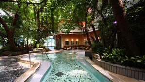 A pool area with a tiled floor, surrounded by trees and plants, and a building with a covered seating area in the background.