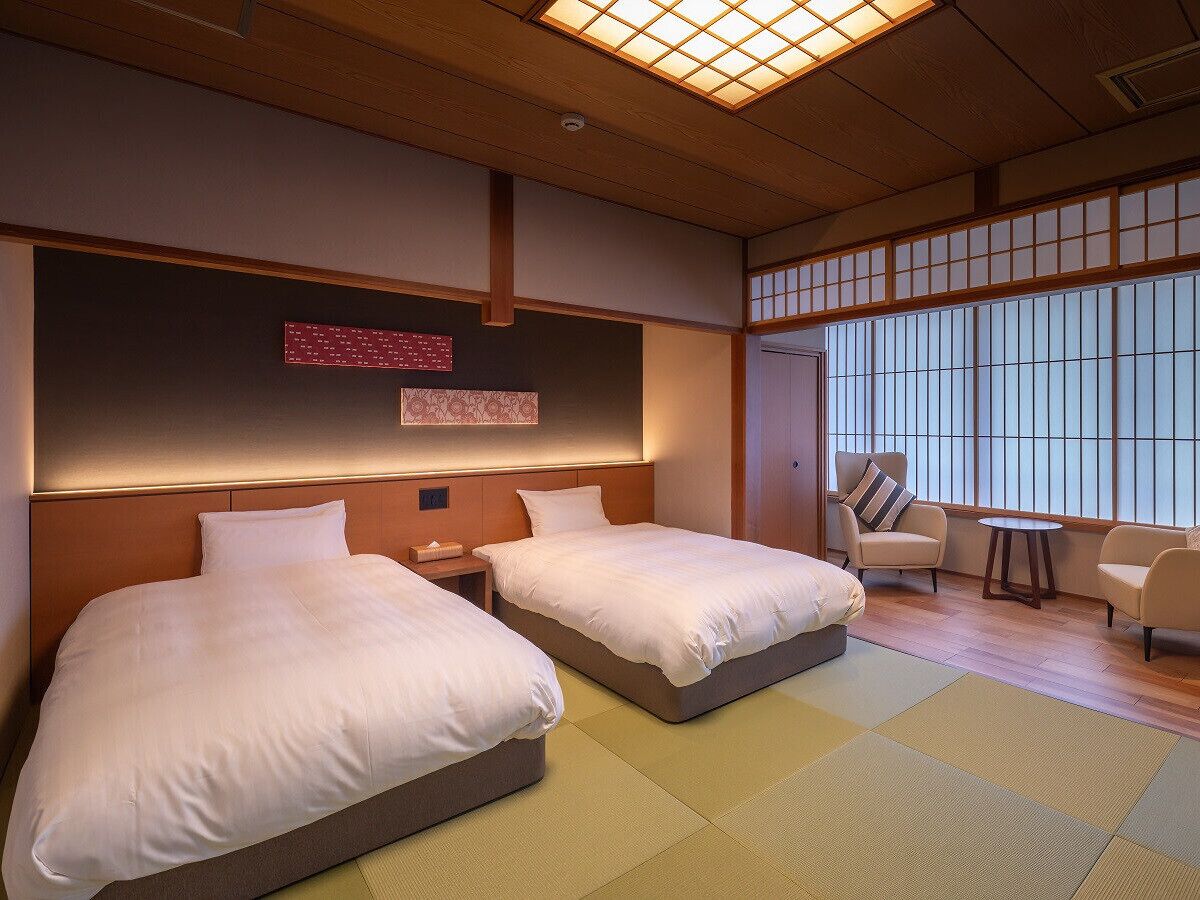 A hotel room with two beds, a sitting area with a chair and a small table, and a large window with a view.