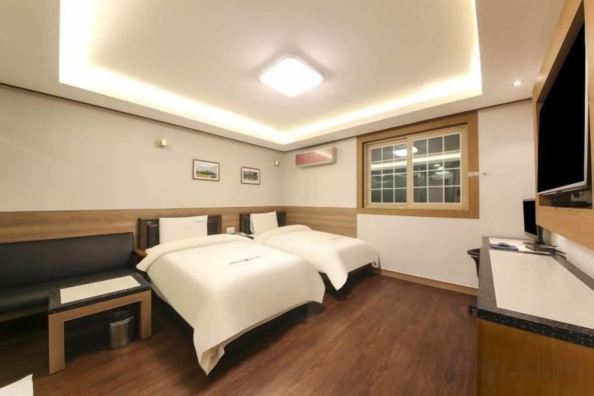 A hotel room with two beds, a bench, a small table, a window, and a sink.