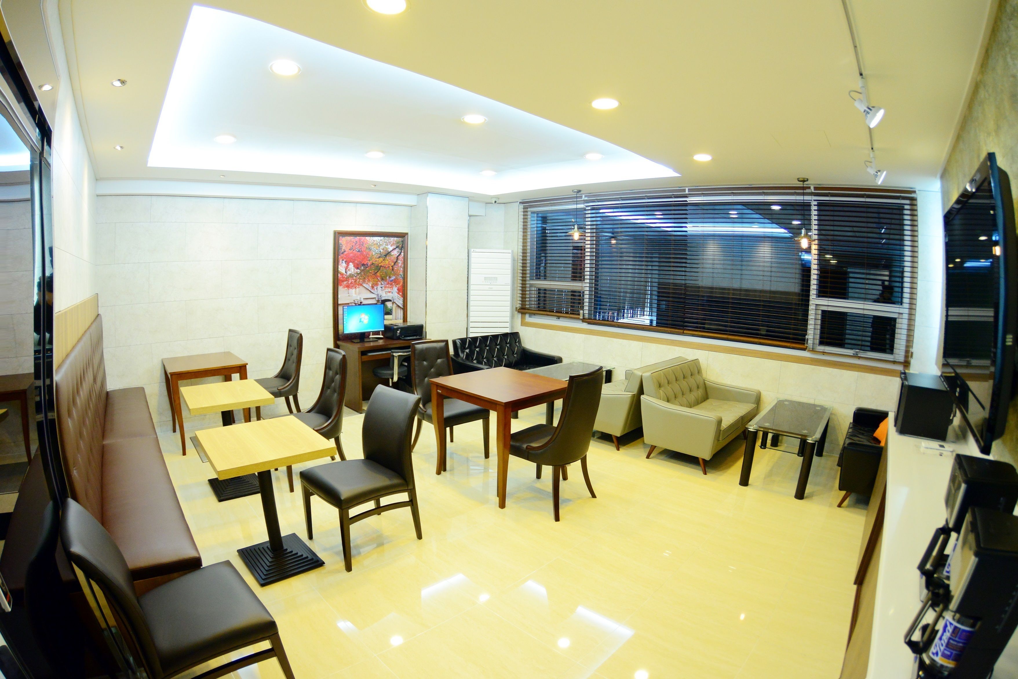 A modern dining area with a high ceiling, recessed lighting, and a large window with blinds. There are multiple seating options including tables, chairs, and a sofa.