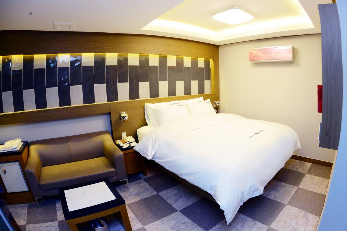 A hotel room with a bed, a sofa, a small table, a nightstand, and a wall-mounted TV.