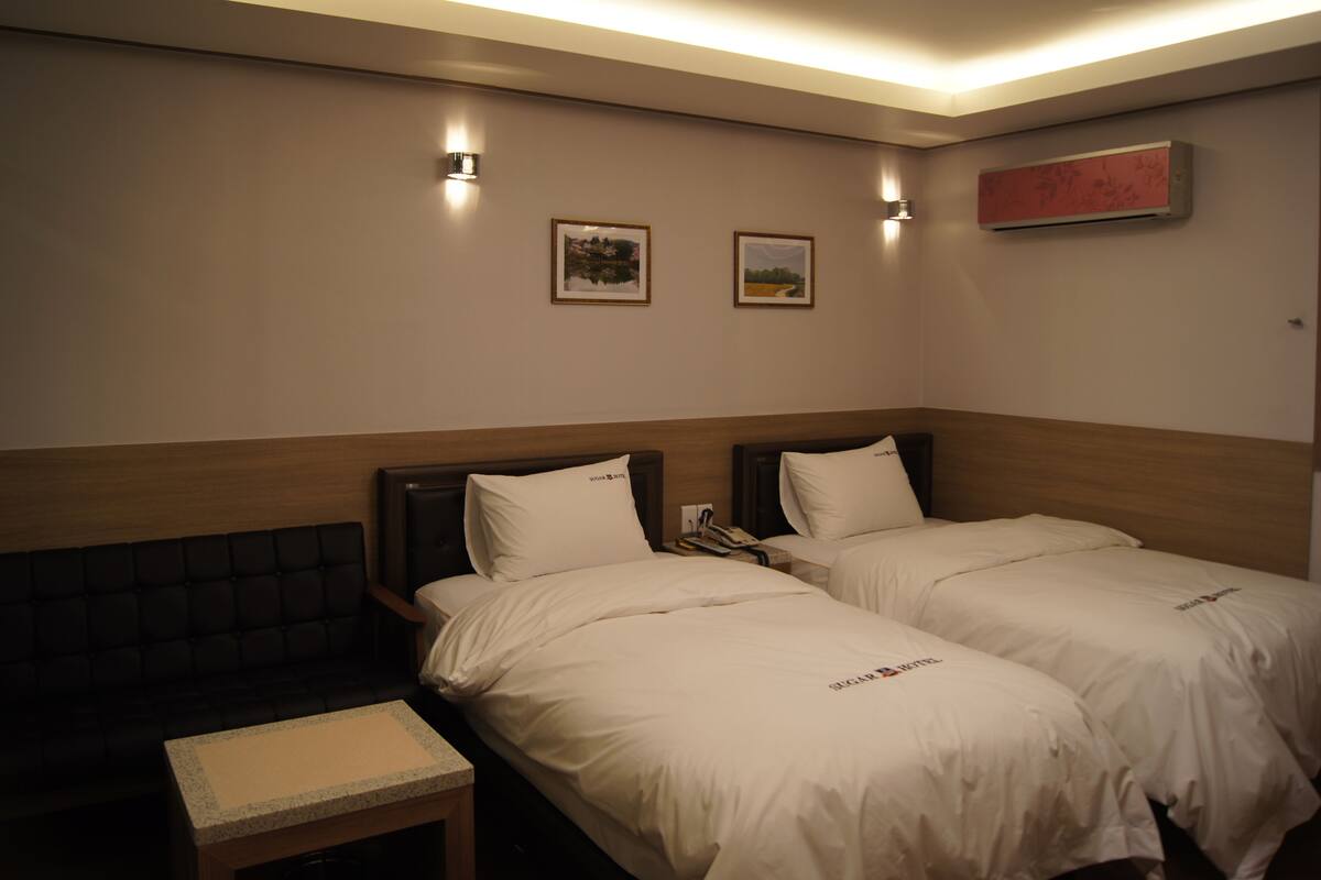 A hotel room with two beds, a small table, and two wall-mounted lights.