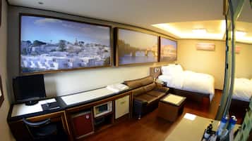 A hotel room with a desk, computer, chair, sofa, bed, and large windows with cityscape views.