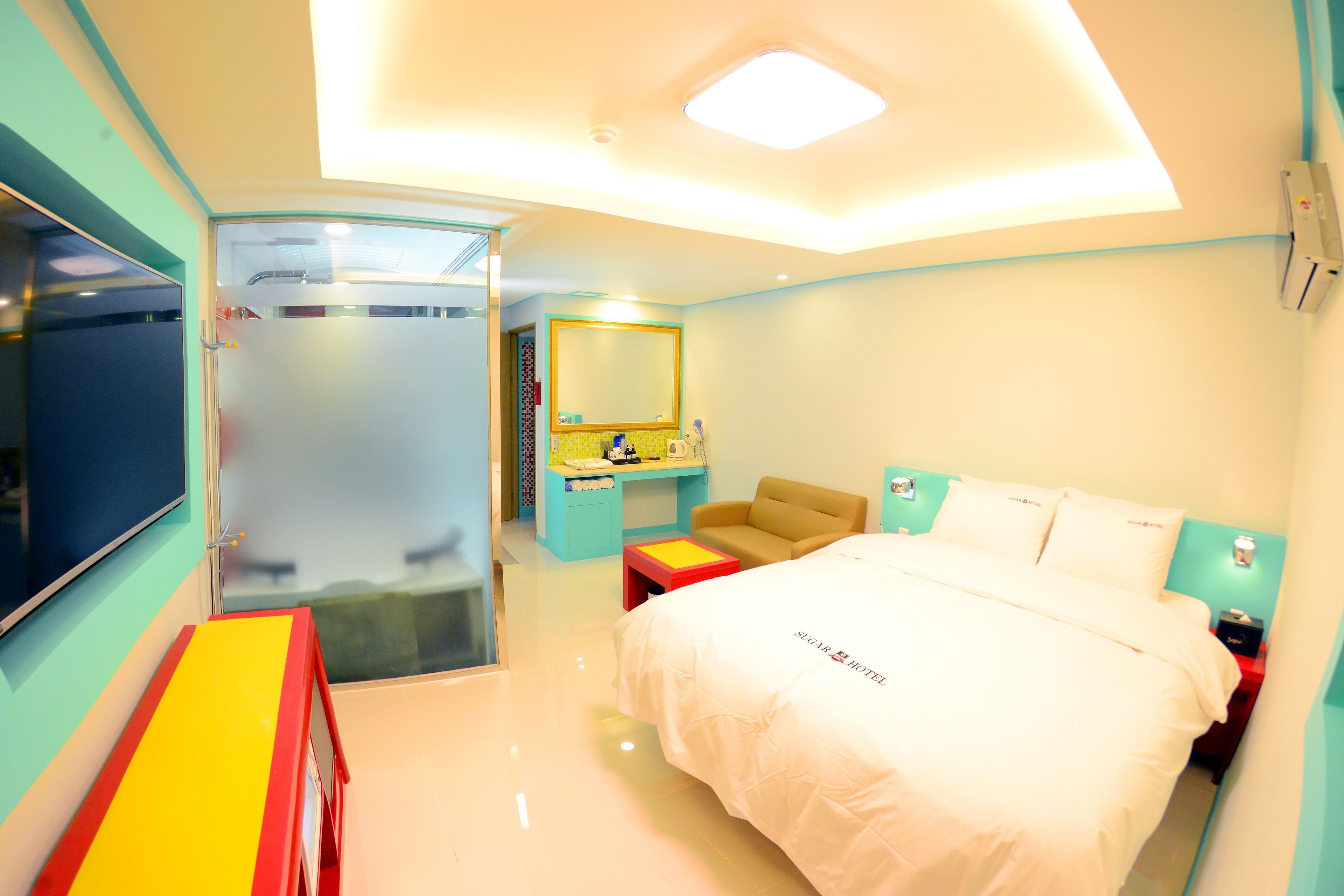 A modern hotel room with a large bed, a desk, a chair, a small table, a mirror, and a bathroom with a glass door.