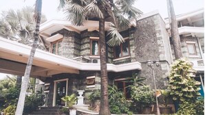 A multi-story building with a stone facade, a central entrance with a white balustrade, and surrounding lush greenery including palm trees. - Arimbi Pejaten Suites (Jakarta)
