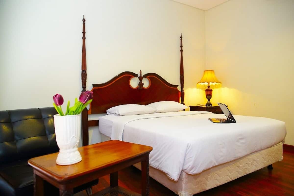 Habitación de hotel con cama con dosel, mesita de noche con lámpara, una silla y un jarrón con flores.