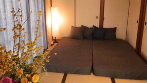 A traditional Japanese-style room with tatami flooring, a futon bed, a bedside lamp, and a floral arrangement. - Daikoku House (Osaka)