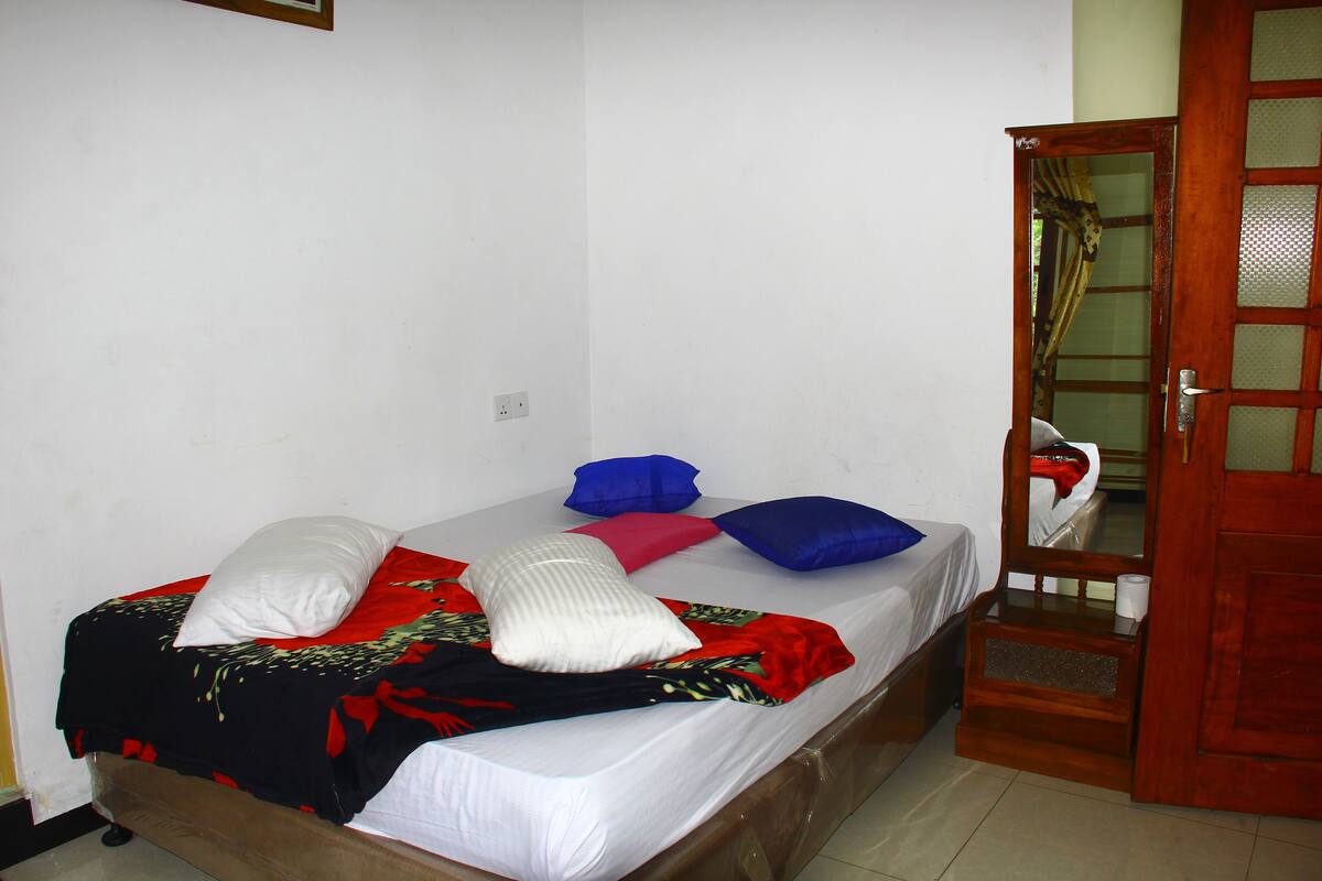 A single bed with a white mattress, red and black blanket, and multiple pillows in white, blue, and pink. A wooden door with glass panels is visible to the right.
