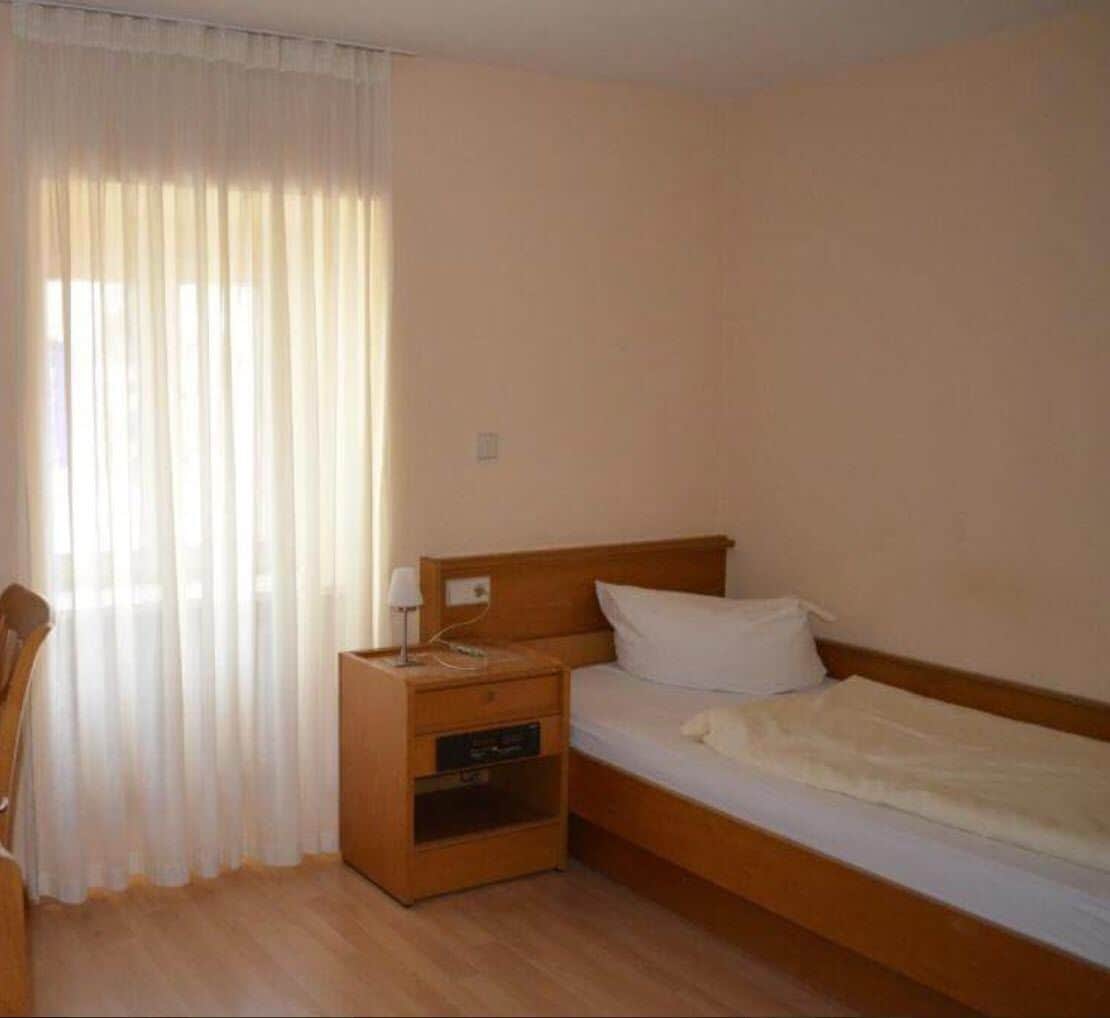 A single bed with a wooden headboard, a nightstand with a lamp, and a window with sheer curtains.