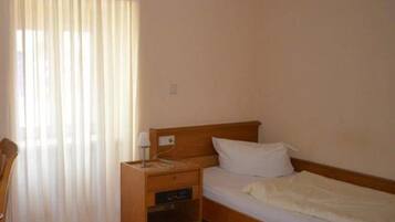 A single bed with a wooden headboard, a nightstand with a lamp, and a window with sheer curtains.