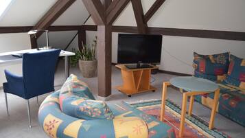 A cozy loft-style room with a high ceiling, exposed wooden beams, a comfortable sofa, a small table, a chair, and a television.