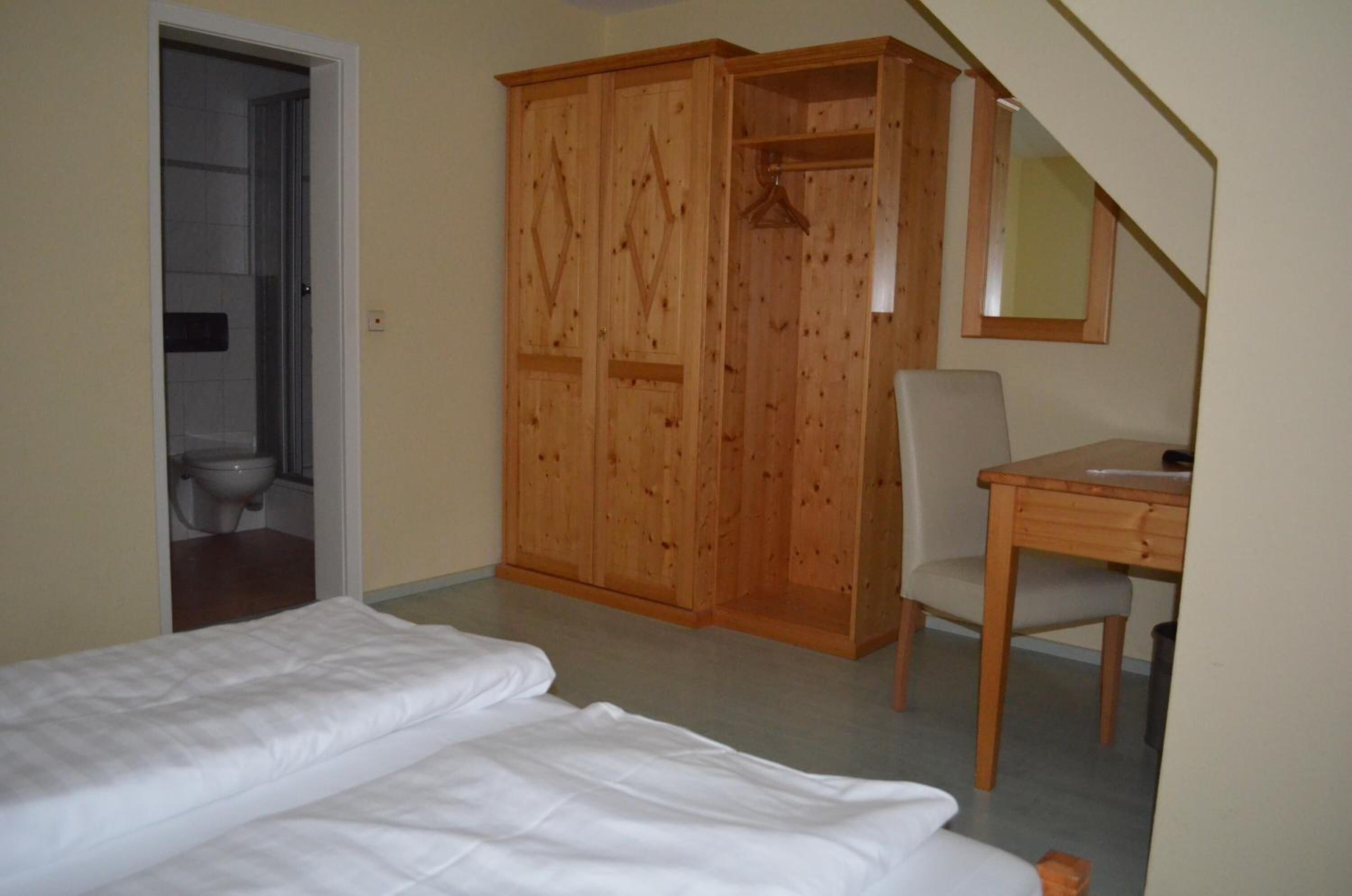 A bedroom with a bed, a wooden wardrobe, a desk, and a chair.