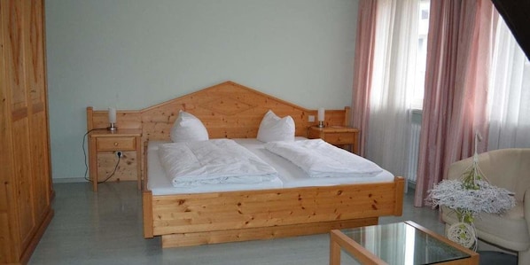 A wooden bed with white linens, a nightstand, a chair, and a glass table in a room with wooden walls and a window with curtains.