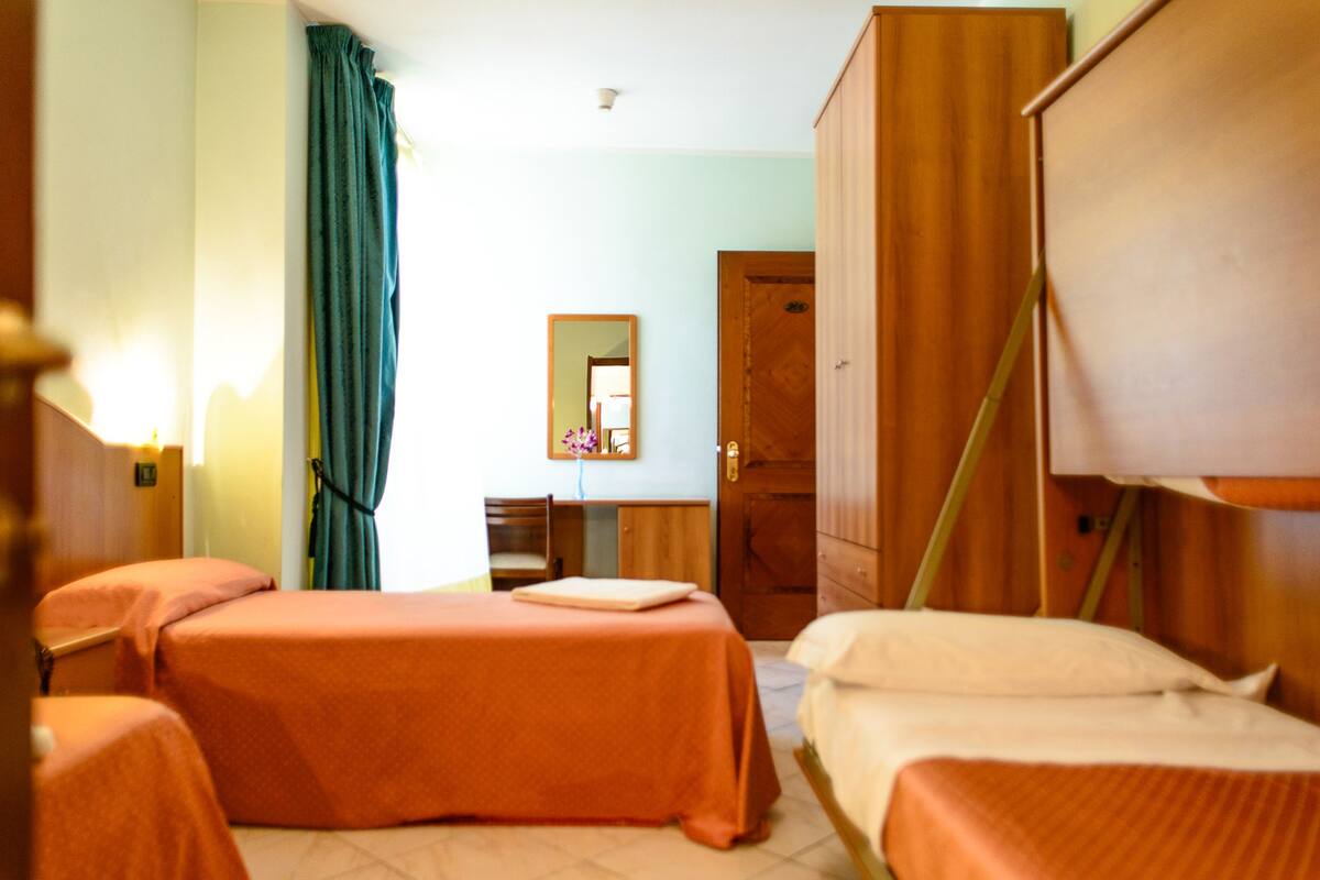 A hotel room with two single beds, a wooden wardrobe, a desk with a chair, a mirror, and a window with curtains.