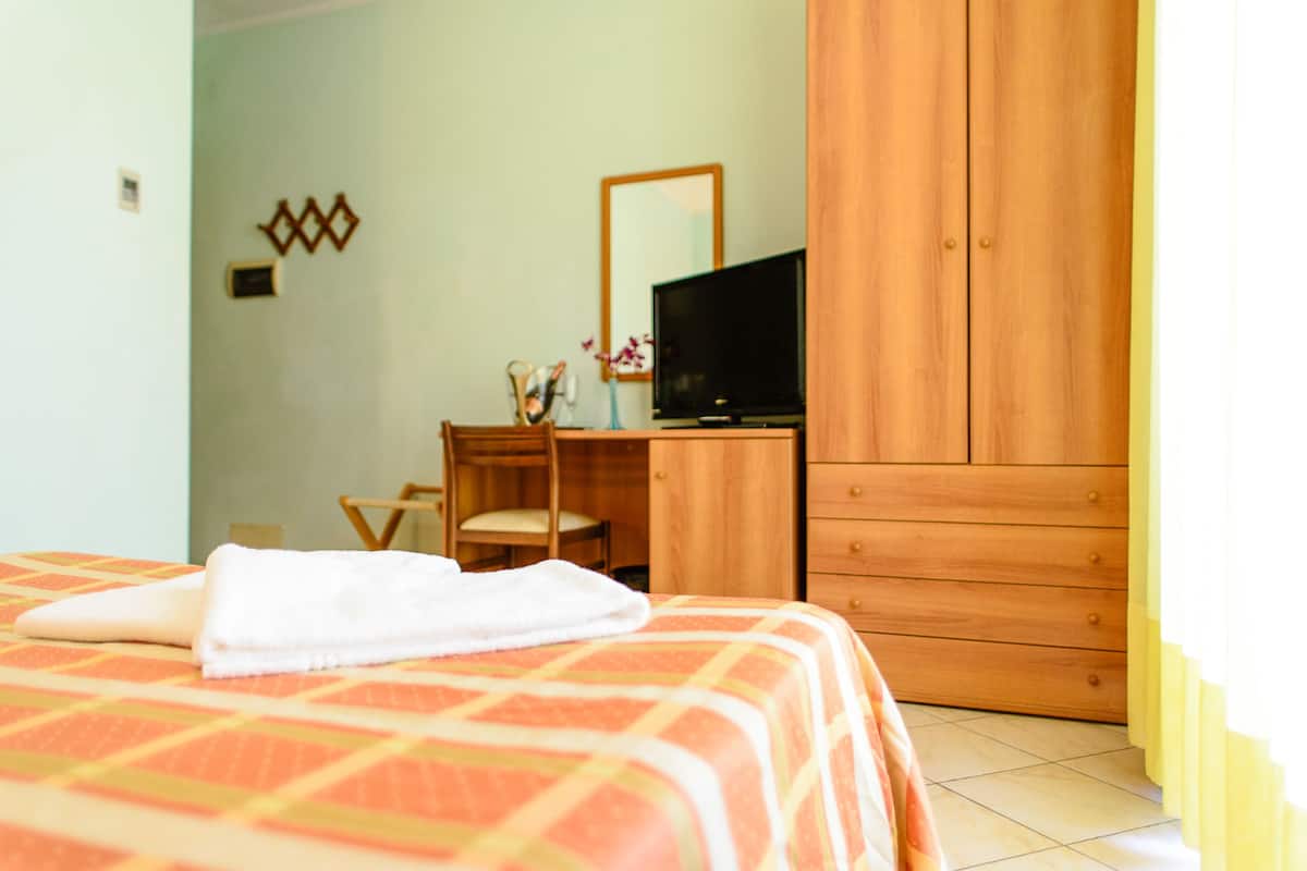 A bedroom with a bed, a wooden wardrobe, a desk with a chair, and a television.