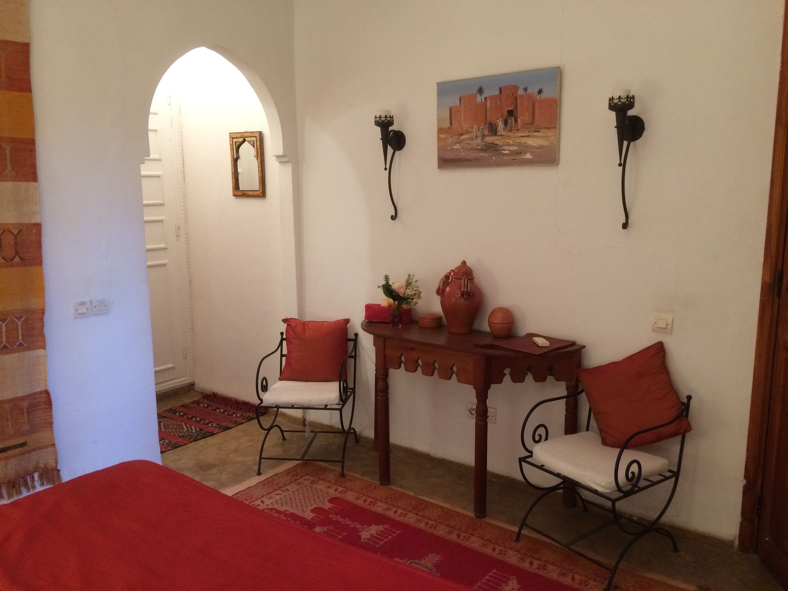 Una habitación con una cama, dos sillas, una mesa con adornos, un cuadro y una lámpara de pared.