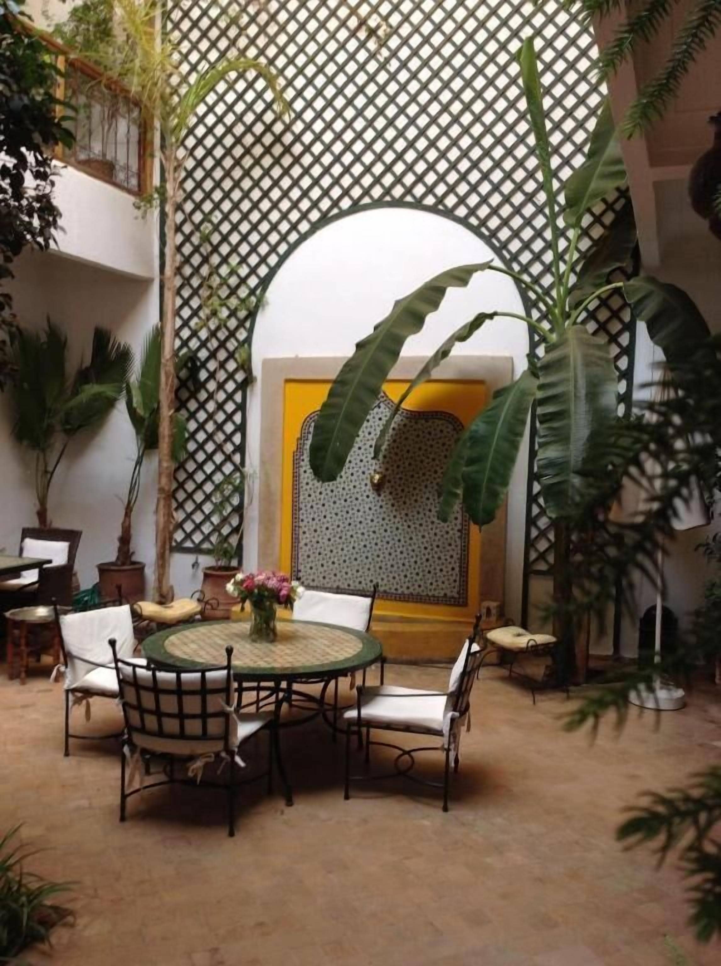 Une table ronde entourée de chaises, située dans une cour, avec des plantes en pot et un mur en treillis.