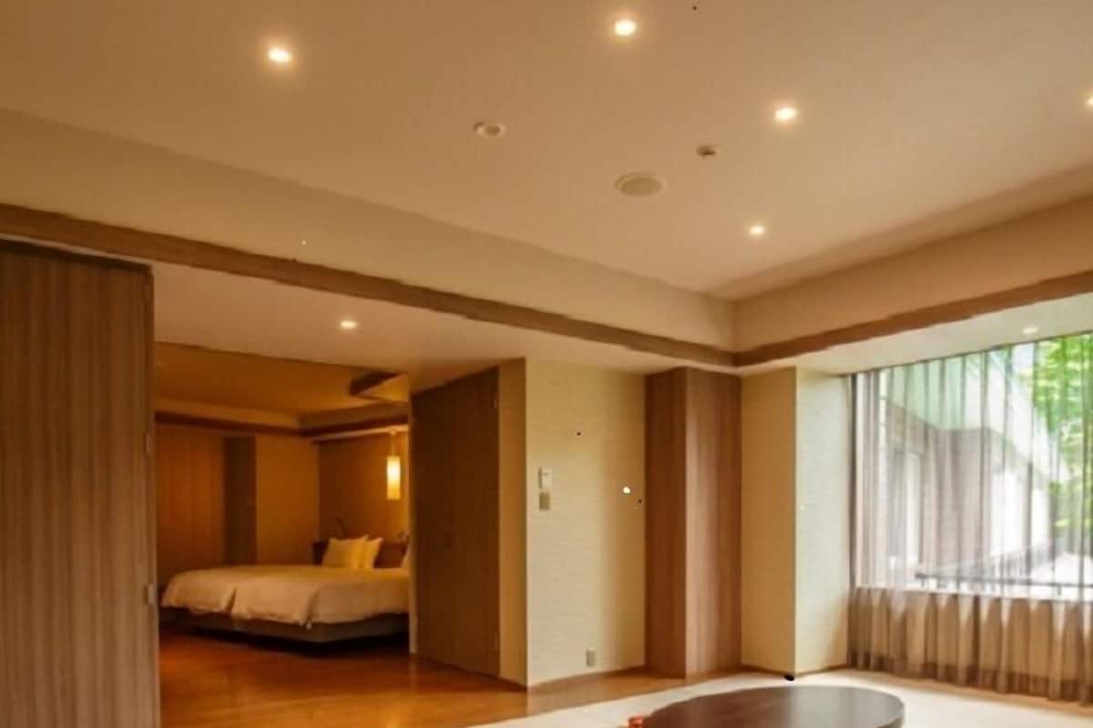 A modern hotel room with a large bed, a dining area with a round table, and a view of greenery outside.